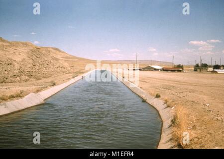 Fotografia di un calcestruzzo rivestito di irrigazione canale di distribuzione passando attraverso il paesaggio arido nel Nuovo Messico, con alcune case in lontananza, un sito relativo al CDC inchiesta del vettore di malattie in acqua risorsa sviluppi, 1976. Immagine cortesia CDC. () Foto Stock