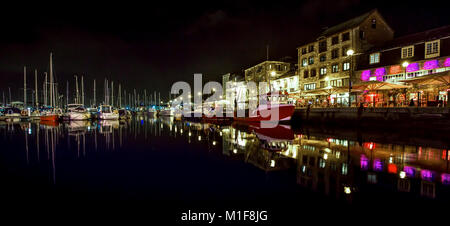 Una vista di varie barche da pesca e da diporto riflessa in mirror come l'acqua con i locali della vita notturna illuminata in background Foto Stock