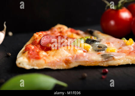 Close up della fetta di italiano Capriciosa pizza di ardesia scura con pomodori e le erbe aromatiche Foto Stock