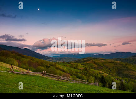 Area rurale al tramonto con la luna sul cielo nuvoloso. bellissimo paesaggio di montagna con campi agricoli e di recinti di legno su pendii erbosi in primavera Foto Stock