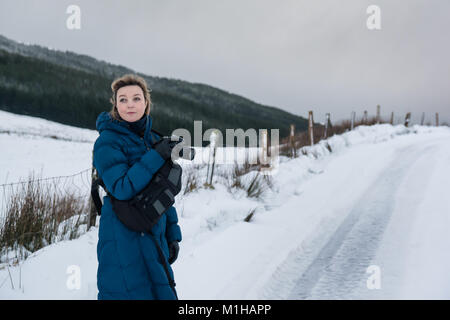 Fotografo femmina in un mantello blu prendendo immagini del paesaggio nella neve in inverno Foto Stock