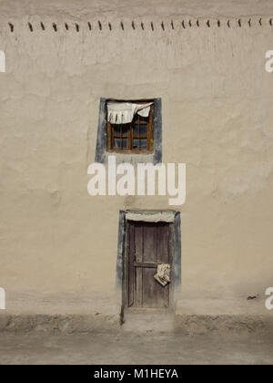 Parete di argilla è una luce di colore beige, con un'antica porta in legno nella parte inferiore e una piccola finestra sulla parte superiore con una tenda bianca. Foto Stock