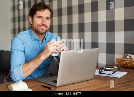 Uomo sorridente in linea di lavoro e di bere il caffè a casa Foto Stock