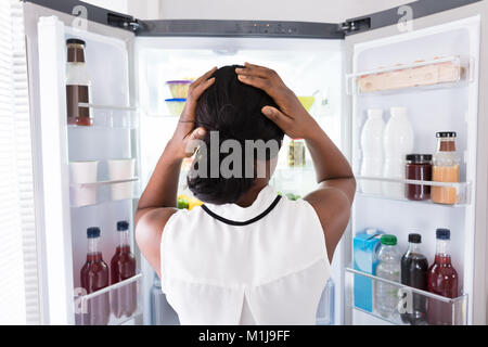 Vista posteriore di un confuso giovane donna africana cercando in frigorifero aperto Foto Stock