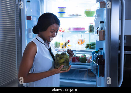 Felice donna africana mangiare Pickle da Jar vicino al frigorifero Foto Stock