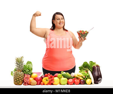 Donna sovrappeso tenendo una ciotola di insalata e flettendo il suo bicipite dietro un tavolo con la frutta e la verdura isolati su sfondo bianco Foto Stock