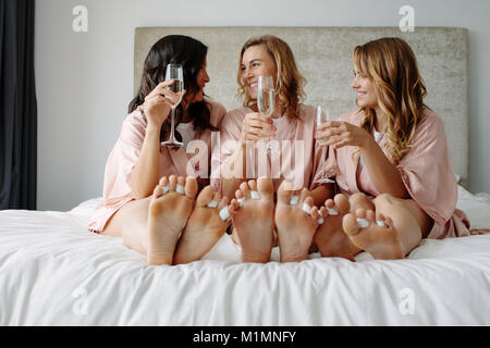 Felice bridesmaids e sposa in vesti di seta seduta sul letto e sorridente. Celebrare la festa di gallina a casa. Foto Stock