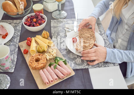 Giovane donna di gustare una sana prima colazione seduti ad un tavolo diffuso con frutta fresca, affettati, formaggio e pane con assortimento di bevande in un Foto Stock