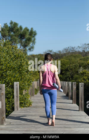 La donna che porta un arrotolato materassino yoga, esterno su un ponte di legno. Vista posteriore Foto Stock