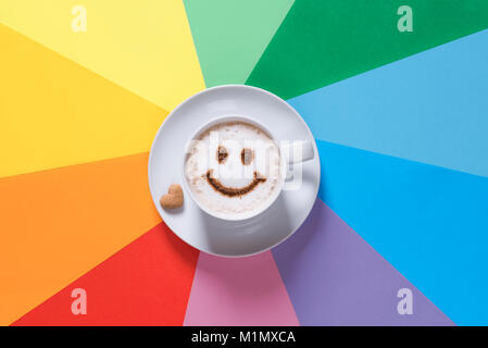 Tazza di cappuccino con una faccina sorridente, sulla schiuma di latte con cacao in polvere e una a forma di cuore nei pressi di zucchero, su un sfondo della carta nei colori dell'arcobaleno. Foto Stock