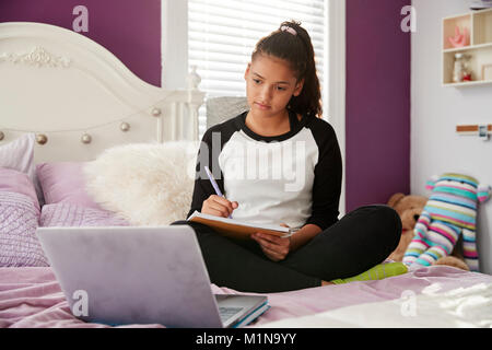 Teen ragazza seduta sul letto a guardare il computer e scrivere note Foto Stock