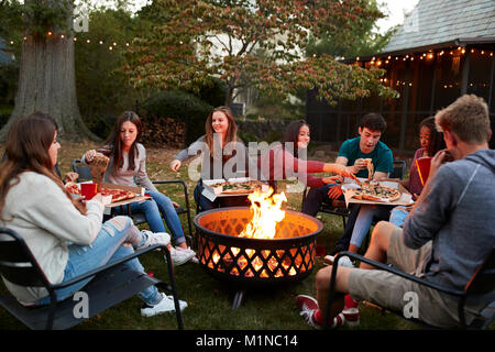 Amici di adolescenti sedersi attorno a una buca per il fuoco mangiare pizze da asporto Foto Stock