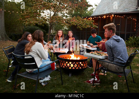 Amici di adolescenti sedersi attorno a una buca per il fuoco mangiare take-away pizza Foto Stock