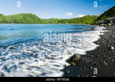Lavare le onde a riva di Pasagshak Bay con il villaggio di Pasagshak in background sul isola di Kodiak in Alaska sudoccidentale. Foto Stock