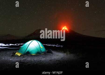 Notte del paesaggio di montagna della penisola di Kamchatka: illuminato campeggio turistico su sfondo eruzione vulcanica, la luce proveniente da proiettori intorno a tenda. La Russia Foto Stock