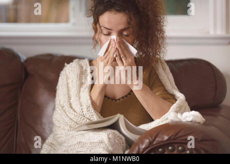 Giovane donna si è ammalato, soffiando il suo naso che cola in un tessuto di carta, di stare a casa con un libro Foto Stock