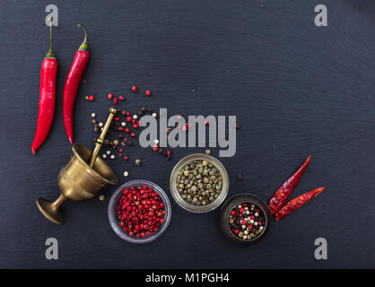Varie grani di pepe e peperoni e pepe in ottone mulino in pietra nera di sfondo, vista dall'alto, spazio di copia Foto Stock