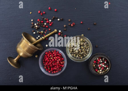 Vari semi di pepe e ottone un mortaio e pestello in pietra nera di sfondo, vista dall'alto, spazio di copia Foto Stock