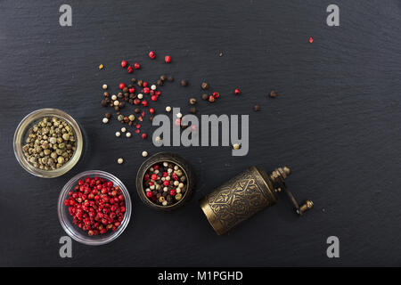 Vari semi di pepe e un pepe in ottone mulino in pietra nera di sfondo, vista dall'alto, spazio di copia Foto Stock