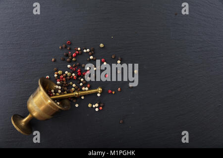 Vari semi di pepe e un mortaio di ottone sulla pietra nera di sfondo, vista dall'alto, spazio di copia Foto Stock