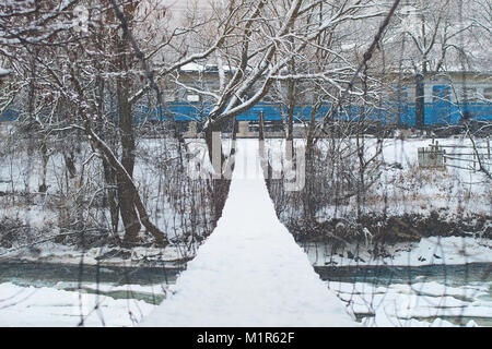 Semplice sospensione ponte corda appesa sopra un fiume di montagna ricoperta di neve con un treno che passa su sfondo inverno Foto Stock