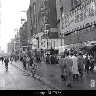 Degli anni Cinquanta, storico, scene di strada, Tokyo, Giappone, uomini in tute impermeabili a piedi lungo un ampio marciapiede esterno passato il Roxy movie theater dopo un recente doccia. Foto Stock