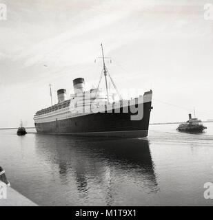 Degli anni Cinquanta, storico rimorchiatori tirare la RMS Queen Elizabeth ocean liner in acque calme di Southampton docks, Inghilterra, Regno Unito. Chiamato dopo la British Regina Madre, è stata la più grande nave passeggeri mai costruito in quel momento (1938) e rimase per 56 anni. Essa è stata utilizzata come un troopship durante il WW2 e solo divenne un ocean liner, come previsto, nel 1946. Foto Stock