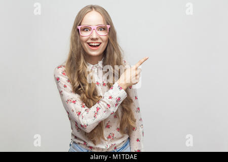 La felicità ragazza, che mostra il dito a copiare lo spazio e guardando la telecamera, con toothy sorridente. Studio shot, isolata su uno sfondo grigio Foto Stock