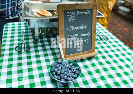 Bocce di frutta da un segno pubblicità Buongiorno pancake con una senza glutine opzione. Persona prepara frittelle in background Foto Stock