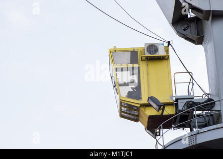 Colpo di giallo cabina di comando con il lavoratore. Vecchie e arrugginite, porta grigia gru di sollevamento carico in nave sul cielo blu chiaro dello sfondo. Attrezzature per la nave di scarico Foto Stock