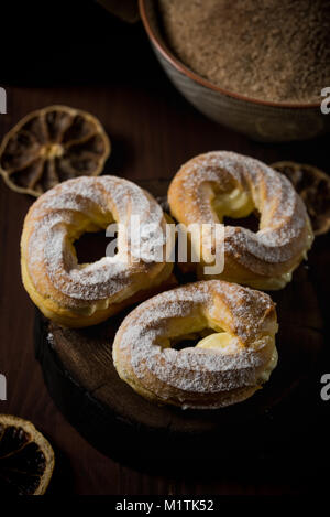 Foto verticale con tre soffi caserecci farciti di crema dolce. I soffi sono coperti da zucchero in polvere. Pochi essiccato anelli arancioni sono versate intorno su d Foto Stock