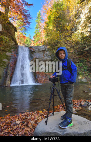 Capretto felice vicino a foto fotocamera sul cavalletto con una cascata in background in autunno Foto Stock