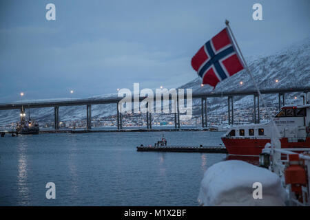 La bandiera norvegese vola nel porto di Tromsø contro Tromsø Bridge (Tromsøbrua) in background a Tromsø in Norvegia durante l'inverno e notte polare. Foto Stock