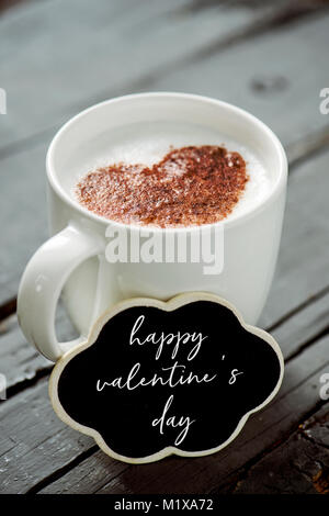 Primo piano di una nuvola a forma di cartello con il testo felice il giorno di San Valentino e una tazza di cappuccino con un cuore disegnato con il cacao in polvere sulla sua schiuma di latte, Foto Stock