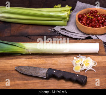 Tagliate a fette le cipolle verdi su una tavola di legno, dietro i grappoli di sedano e marinato pomodori ciliegia in una ciotola Foto Stock