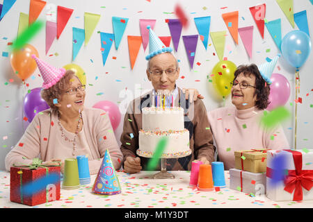 Uomo anziano con due donne anziane soffiando candele su una torta di compleanno contro una parete con decorazione di bandiere e di palloncini Foto Stock