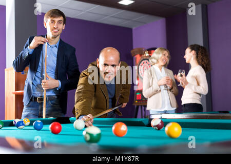 Gruppo di amici di età diversa giocando a biliardo e sorridente in biliardo club. Focus sull'uomo maturo Foto Stock