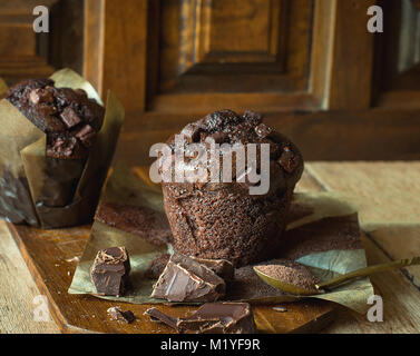 Deliziosa casa cotto scaglie di cioccolato muffin in carta marrone sul tagliere di legno. Cacao in cucchiaio. Cucina rustica interni in stile rurale. Siz quadrato Foto Stock