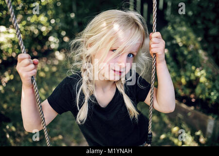 Adorabili poco ragazza bionda giocando da se stessa su un albero oscillante nel suo cortile in una giornata di sole Foto Stock