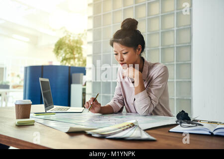 Giovane architetto asiatico seduto da solo ad una tabella in un ufficio moderno lavorando su una progettazione edilizia Foto Stock