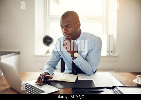 Giovane africano business executive che indossa una camicia e cravatta seduto alla sua scrivania in un ufficio a lavorare online con un computer portatile Foto Stock