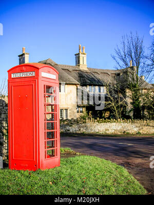 Un telefono rosso scatola in un villaggio con cottage con il tetto di paglia dietro Foto Stock