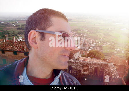 Closeup profilo di mezza età uomo caucasico con occhiali da sole contemplando da sopra un vecchio villaggio italiano al tramonto. Foto Stock