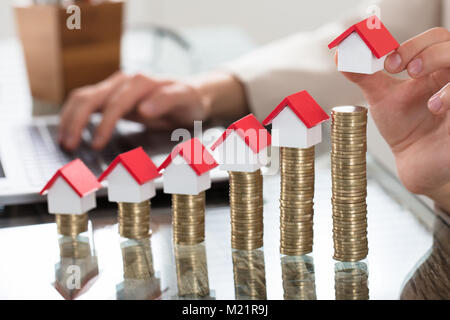 Case in miniatura oltre la crescente pila di monete sul banco di vetro al posto di lavoro Foto Stock