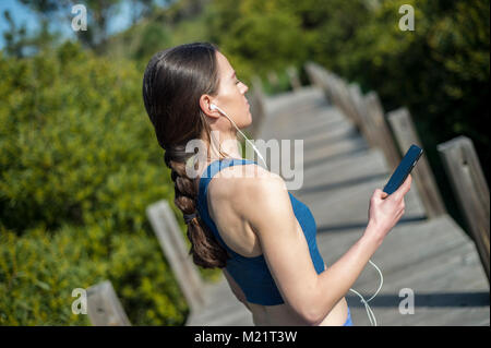 Donna che indossa un reggiseno sportivo ascolta musica sul suo smart phone durante l esercizio al di fuori nel sole Foto Stock