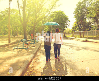 Migliore Femmina degli amici condividendo un ombrello mentre si cammina insieme in un parco con una forte luce del sole durante il giorno Foto Stock