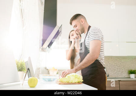 Vista laterale ritratto di giocoso coppia giovane cooking per colazione in accogliente cucina moderna, illuminate dalla luce del sole, spazio di copia Foto Stock