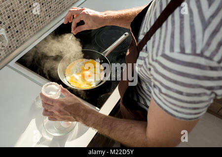 Angolo alto close up dei maschi di mani salatura delle uova in padella durante la cottura prima colazione nella cucina moderna illuminate dalla luce del sole Foto Stock