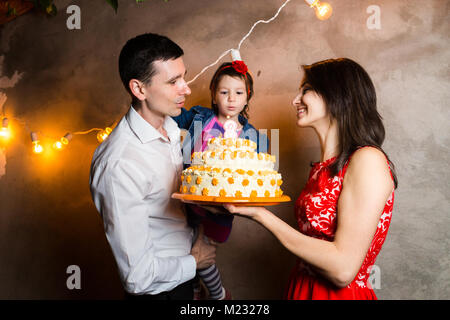 Tema famiglia compleanno per bambini e soffiando le candeline sulla torta di grandi dimensioni. giovane famiglia di tre persone in piedi e azienda figlia di 5 anni ho Foto Stock