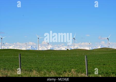 Una fattoria di turbine eoliche a sedersi in un campo di erba verde con una vista incredibile della coperta di neve montagne rocciose della distanza Foto Stock